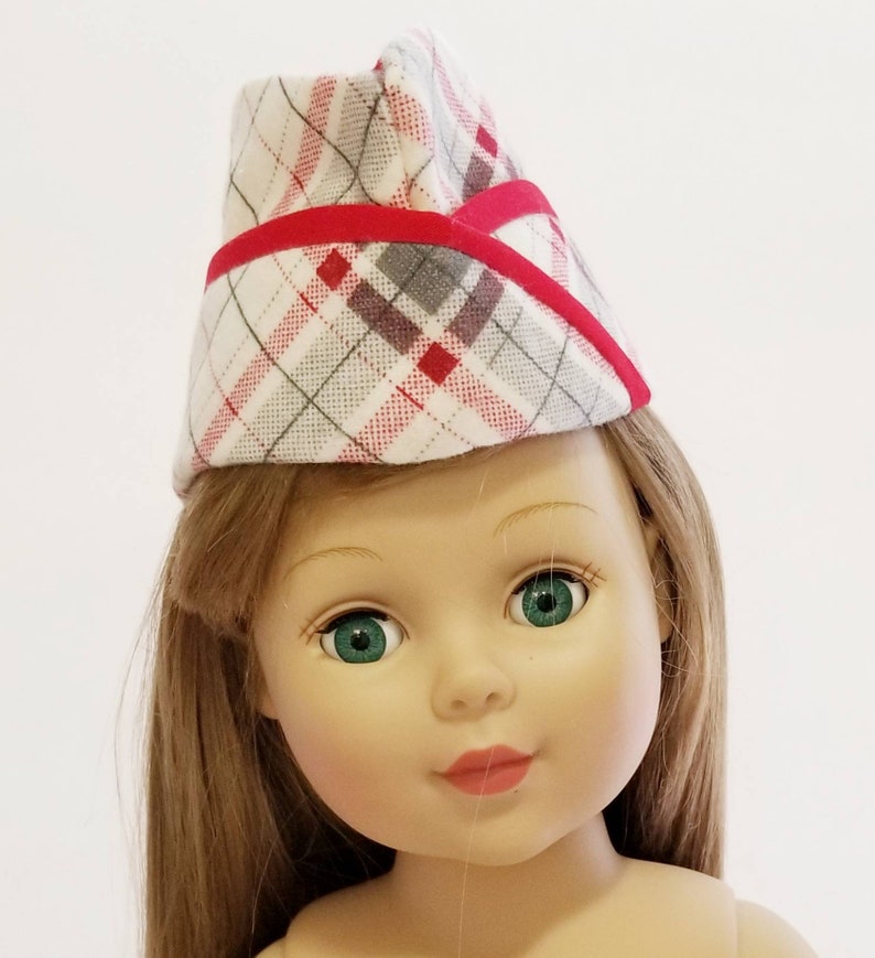 K&ouml;nnte beinhalten: Eine Puppe mit einem rot-grauen karierten Hut mit rotem Besatz. Die Puppe hat lange braune Haare und gr&uuml;ne Augen.