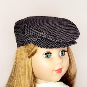 May include: A dark gray houndstooth pattern newsboy cap. The cap is made of a textured fabric and features a rounded crown and a small, stiff brim. The cap is designed for a doll.