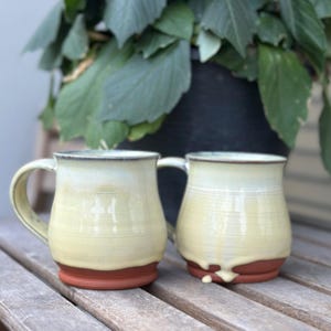May include: Two handmade ceramic mugs with a cream glaze and a reddish-brown base. The mugs have a simple, rounded shape with a handle. They are on a wooden surface. A large green plant is in the background.