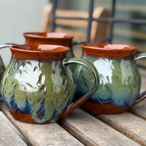 Puede incluir: Tazas de cerámica hechas a mano con un esmalte texturizado verde y azul, acentuadas por un borde y asa marrones. Las tazas están sobre una superficie de madera, mostrando su diseño artesanal único. Ideales para bebidas calientes.