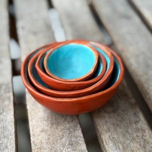 Puede incluir: Un juego de cuatro cuencos de cerámica hechos a mano, anidados entre sí. El exterior es de un cálido marrón rojizo, mientras que el interior de cada cuenco es de un vibrante azul turquesa. Los cuencos están dispuestos sobre una superficie de madera envejecida.