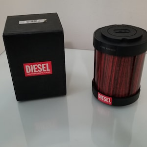Może przedstawiać: Czarny, cylindryczny odświeżacz powietrza z czerwonym filtrem i napisem "DIESEL" nadrukowanym na boku. Odświeżacz powietrza jest zapakowany w czarne pudełko z napisem "DIESEL" nadrukowanym z przodu.