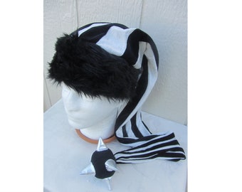 Può includere: Cappello a strisce bianche e nere con bordino in pelliccia nera e coda a strisce bianche e nere. Il cappello è su una testa di manichino bianca. C'è una palla nera con punte e dettagli argentati sulla superficie bianca.