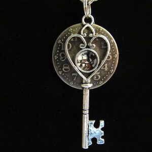 May include: A silver pendant necklace with a key design. The key features a circular charm with a heart-shaped frame and a small image at the centre. The charm has numbers around the edge, and the key has a detailed, ornate design.
