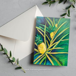 Könnte beinhalten: Grußkarte mit einem Gemälde von Ananas mit grünen Blättern und einem dunkelblauen Hintergrund. Ein weißer Umschlag liegt daneben. Die Website-Adresse www.mariasingletonart.com ist unten aufgedruckt.