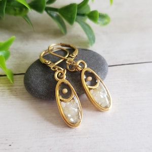 May include: Gold-toned oval earrings with a clear resin center filled with small, white, irregular pieces. The earrings have a small, round, gold-colored accent near the top and a lever-back closure. The earrings are displayed on a gray stone.