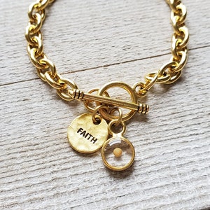 May include: Gold-tone chain bracelet with a toggle clasp. The bracelet features two charms: a round charm with the word "FAITH" and a clear charm containing a mustard seed. The bracelet is displayed on a light wood surface.