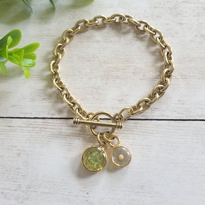 May include: Gold-toned chain bracelet with a toggle clasp. The bracelet features two small, round charms: one is a light green color, and the other is a pale gray. The bracelet is displayed on a white wooden surface.