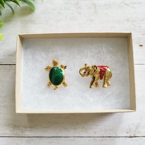 May include: Two gold-toned brooches: a turtle with a green center and an elephant with a red and gold blanket. The brooches are displayed in a small, open cardboard box with white padding.