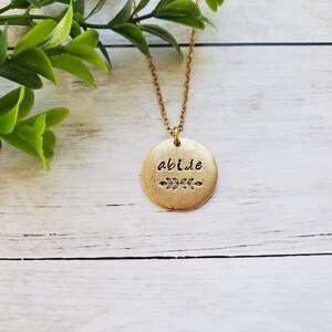 May include: Gold necklace with a round pendant that says "abide" with a small leaf design.