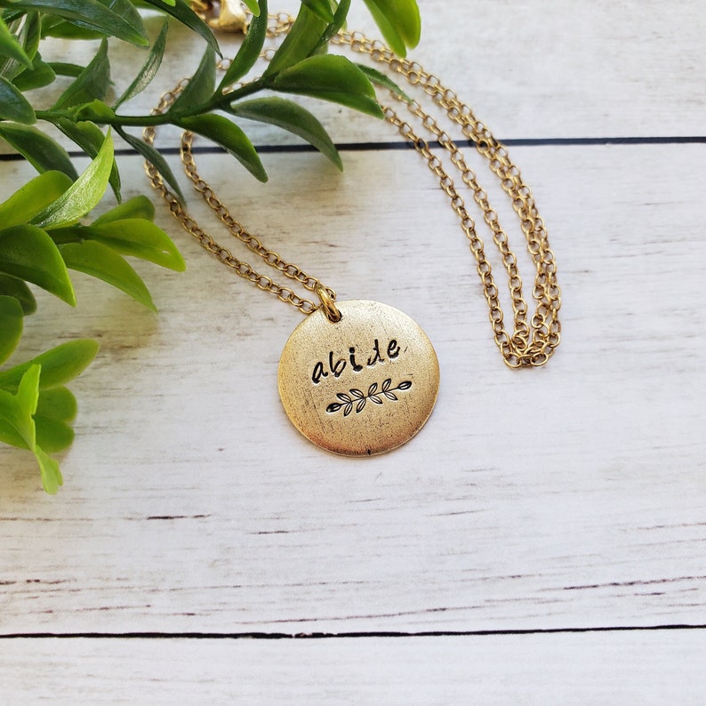 May include: Gold necklace with a round pendant that says "abide" with a small leaf design underneath the word.