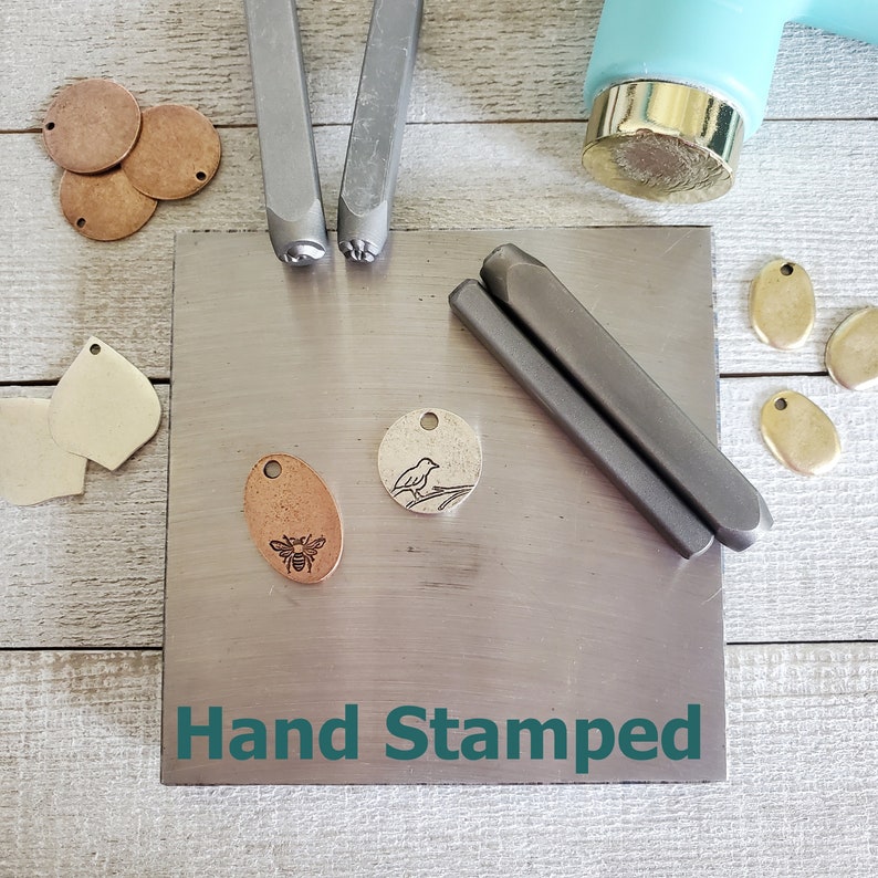 May include: A metal surface with the words "Hand Stamped" printed on it. Two metal stamps are on the surface, one with a bird design and the other with a bee design. Two metal charms are on the surface, one with a bird design and the other with a bee design.