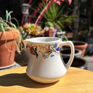 May include: A small, white ceramic creamer with a curved handle and spout. The creamer is decorated with a floral and bird design in shades of orange, blue, and green. The creamer sits on a wooden surface, with a blurred background of plants and pots.