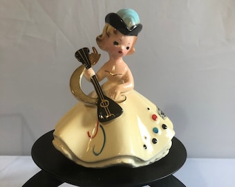 May include: A vintage ceramic figurine of a woman in a yellow dress, playing a black and gold instrument. She wears a blue and black hat. The figurine sits on a black metal stand.