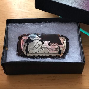 May include: A rectangular, brown and silver brooch with an intricate design of figures and vases, presented in a black gift box with white padding. The brooch has a polished finish and is likely made of metal.