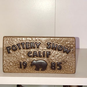 May include: A rectangular ceramic plaque with the words "POTTERY SHOW CALIF 1985" embossed in dark brown. A bear silhouette is centered below the word "CALIF". The plaque has a textured, speckled, tan background.