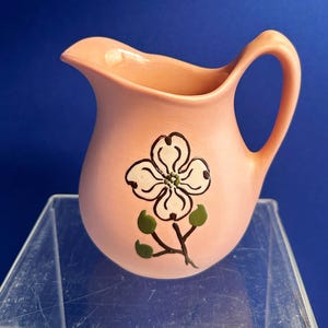 May include: A small, pink ceramic pitcher with a handle and spout. The pitcher is decorated with a white and black floral design with green leaves. The background is a solid blue.