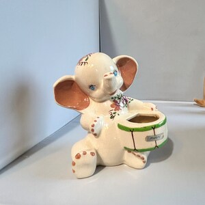 May include: A ceramic elephant figurine in cream with pink ears and floral accents. The elephant holds a green drum with the word "PEANUTS" on it. The figurine has blue eyes and brown spots on its feet.