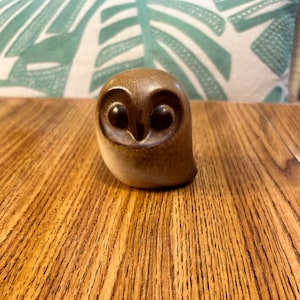 May include: A small, brown ceramic owl figurine with large, dark eyes. The owl has a smooth, rounded body and is sitting on a wooden surface. The background includes a pillow with a green and white leaf pattern.