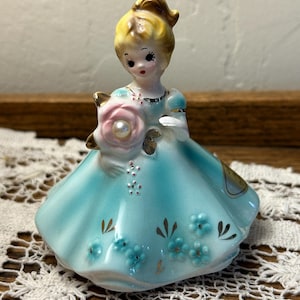 May include: A ceramic figurine of a person with blonde hair wearing a light blue dress with gold accents. The figure holds a large pink rose with a central pearl accent. The dress features small blue floral details and gold leaf patterns.