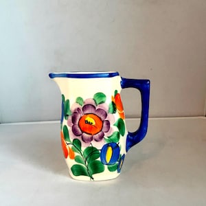 May include: A ceramic pitcher with a white base, adorned with a vibrant floral design in purple, orange, and blue. The handle and rim are a deep blue. The pitcher has a spout for easy pouring.
