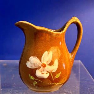 May include: A small, brown ceramic pitcher with a white flower design. The pitcher has a curved handle and a spout for pouring. The flower has a yellow center and green leaves. The pitcher is likely used for serving liquids.