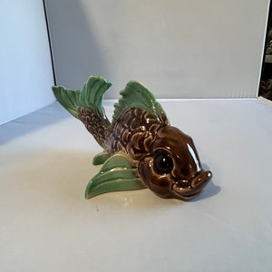 May include: A ceramic fish figurine with a brown body, green fins, and large black eyes. The fish has a detailed scale pattern and is posed as if swimming. The figurine is a decorative item.