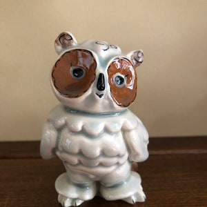 May include: A ceramic owl figurine with a light blue body, brown eyes, and brown accents. The owl has a round face and a slightly ruffled appearance. The figurine is standing on a wooden surface.
