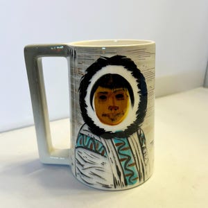 May include: A ceramic mug with a square-shaped handle featuring a hand-painted illustration of a person wearing a dark fur-trimmed hood. The design includes white, black, brown, and turquoise colors with linear etched details on the mug surface.