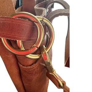 May include: Close-up of a brown leather bag with a brass clasp and strap. The strap is attached to the bag with a metal ring.
