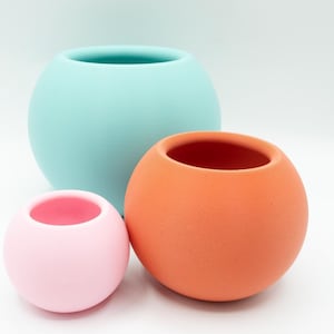 May include: Three round ceramic planters in different sizes. The largest planter is light blue, the middle planter is orange, and the smallest planter is pink.