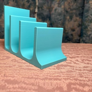 May include: A turquoise desk organizer with four curved slots. The organizer is made of a solid material and sits on a patterned surface. The design is modern and functional for office or home use.