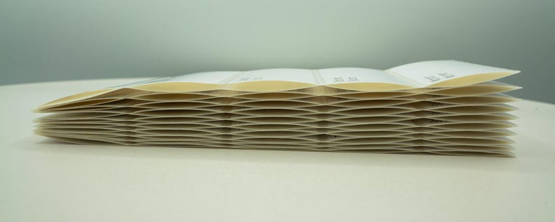 May include: A stack of white paper labels with a slightly yellowed tint. The labels are folded in a zig-zag pattern, creating a wave-like effect.