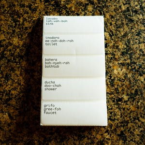 May include: A set of five white labels with black text, each with a Spanish word and its English translation. The words are "lavabo" (sink), "inodoro" (toilet), "ba&ntilde;era" (bathtub), "ducha" (shower), and "grifo" (faucet).