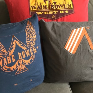 May include: Three decorative throw pillows with different designs. The top pillow is red with a brown and yellow graphic that says "Wade Bowen West 84". The middle pillow is blue with a black and orange graphic of a winged ace of spades with the text "Wade Bowen". The bottom pillow is gray with an orange and white striped graphic.