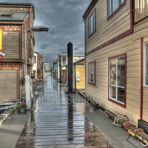 Puede incluir: Una pasarela estrecha de madera bordeada de casas coloridas a ambos lados. La pasarela está mojada por la lluvia reciente, reflejando el cielo y los edificios. Las casas son de madera y tienen una variedad de colores, incluyendo amarillo, rojo y azul.