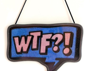 May include: A sign in the shape of a speech bubble with the letters "WTF?!" in pink glitter. The background is blue with a black border, and the sign hangs from a black string.