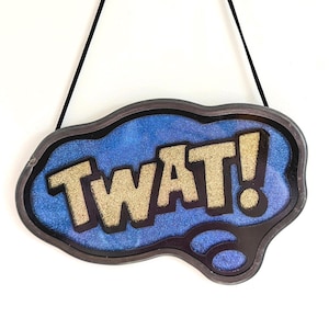 May include: A decorative sign with the word "TWAT!" in gold glitter letters. The sign has a blue background with glitter and a dark brown border. It hangs from a black string.