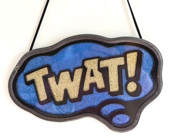May include: A decorative sign with the word "TWAT!" in gold glitter letters. The sign has a blue background with glitter and a dark brown border. It hangs from a black string.