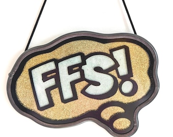 May include: A decorative sign with the text "FFS!" in white letters. The sign has a gold glitter background and a dark brown border. It is hanging from a black string against a white background.