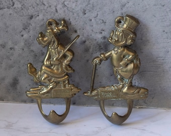 May include: Two brass wall hooks featuring cartoon characters. The hook on the left features a dog-like character holding a stick. The hook on the right features a duck-like character holding a golf club. Both hooks have a single hook at the bottom.