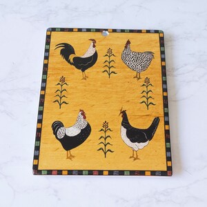 May include: A yellow wooden cutting board with a black and white rooster and hens design. The board has a black, white, red, green, and blue border.