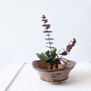 May include: A brown ceramic vase with a wide, flared opening and a rounded base. The vase has a rustic, hand-thrown look and is decorated with brown and white glaze. The vase is filled with a small arrangement of green and purple foliage.