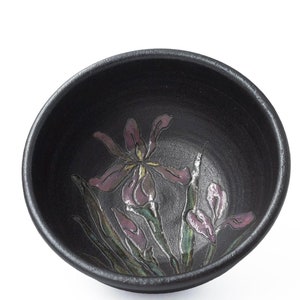 May include: A black ceramic bowl with a floral design. The bowl features a pink and green iris flower with a yellow center.