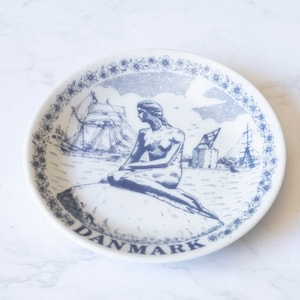 May include: A white ceramic plate with a blue and white design featuring the Little Mermaid, a ship, and a crane. The plate has a blue border with a floral pattern and the word "DANMARK" written in blue at the bottom.