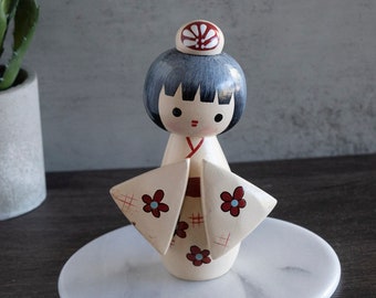 May include: A wooden doll with a white and red kimono, black hair, and a red and white hat. The doll is standing on a white marble base.