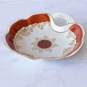 May include: A white ceramic dish with a red rim and gold floral design. The dish has a handle on the side and a small red circle in the center.