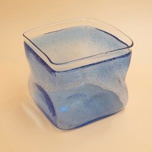 May include: A square, light blue glass vase with a textured surface. The vase has a slightly curved shape and a clear rim. The vase is likely used for decorative purposes.