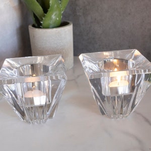May include: Two clear glass candle holders with a geometric design. Each holder has a square base and a pointed top, creating a diamond shape. The holders are filled with tealight candles.