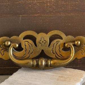 May include: A decorative brass drawer pull with a floral design. The pull has a loop handle and a decorative backplate.